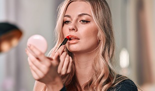 A young woman holding a mirror, applying red lipstick.