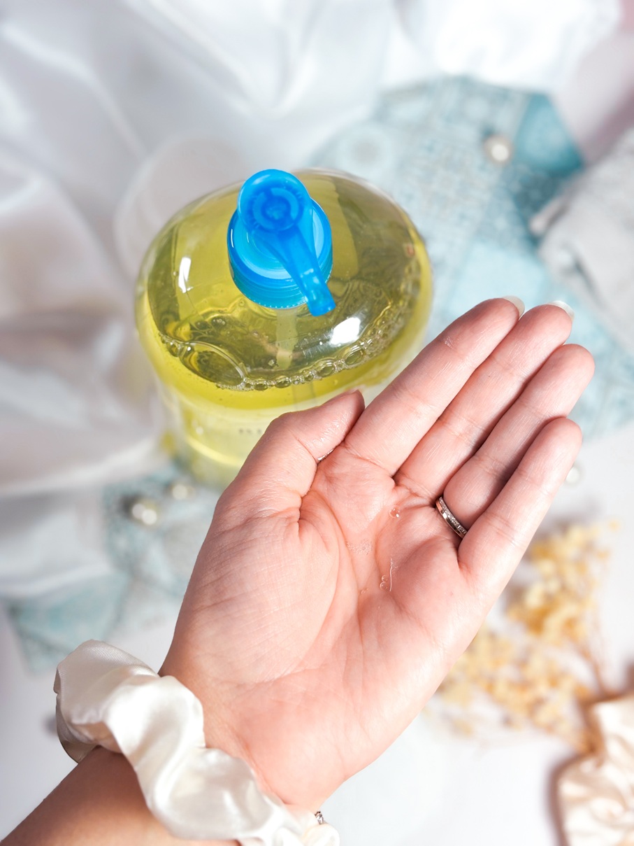 Main montrant une noisette de Bioderma Atoderm Huile de Douche dans la paume, avec le flacon en arrière-plan sur un fond soigné avec tissu satiné, perles, fleurs séchées et chouchou satiné.