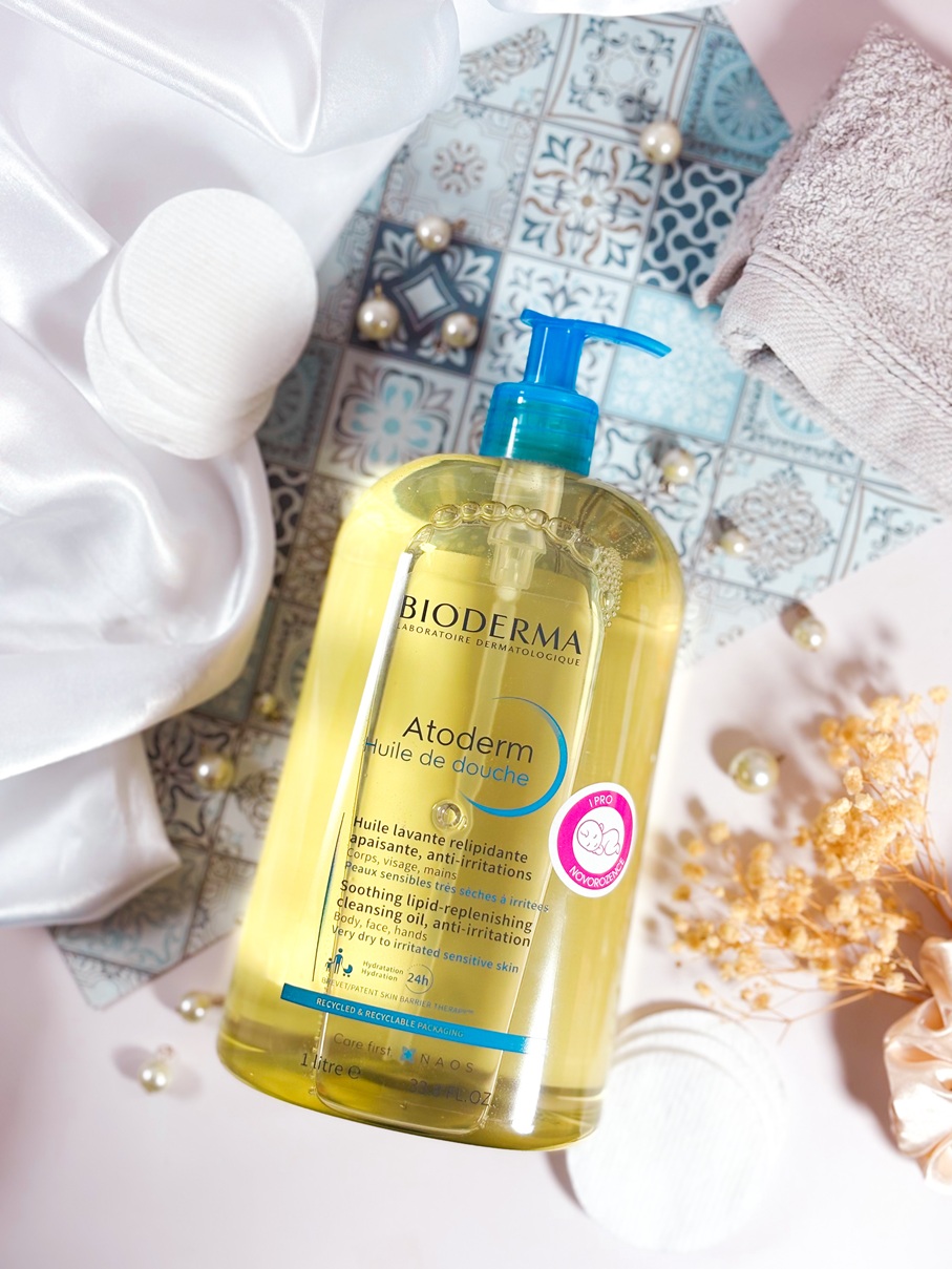 Le gel lavant Bioderma Atoderm Huile de Douche posé sur un fond soigné avec tissu satiné, perles, fleurs séchées, serviette et accessoire de bain.