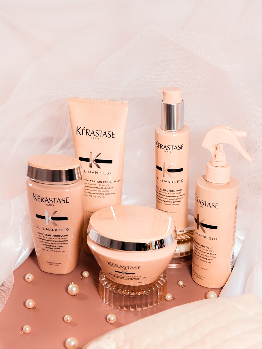 La gamme Kérastase Curl Manifesto réunie sur un fond délicat avec voile en tulle et perles, composée de plusieurs soins pour cheveux bouclés dans leurs flacons et pots rose nude.