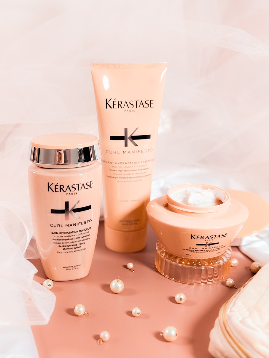 Les soins Kérastase Curl Manifesto, dont shampoing, après-shampoing et masque ouvert, posés sur un fond délicat avec voile en tulle et perles.