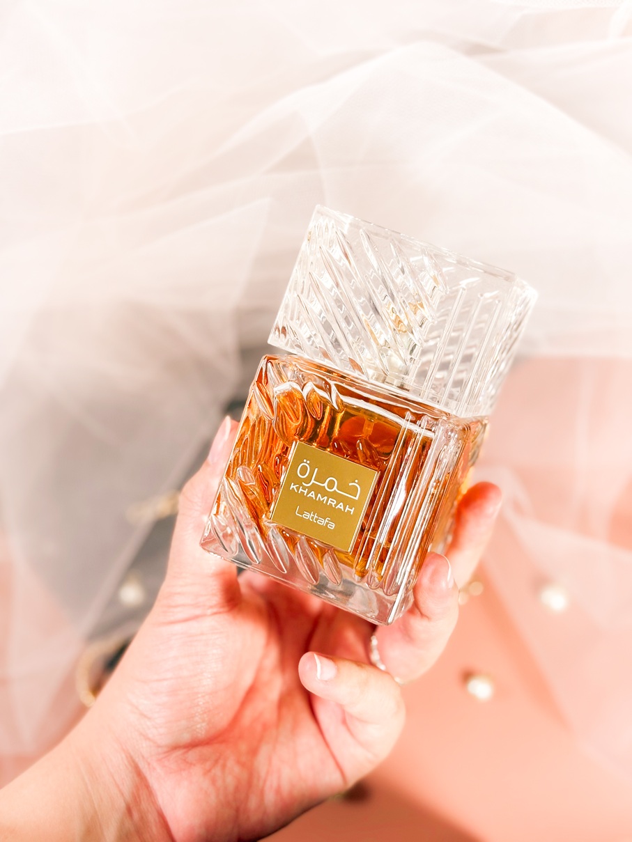 Main tenant le parfum Lattafa Khamrah dans son flacon facetté ambré, sur un fond délicat avec voile en tulle et perles.