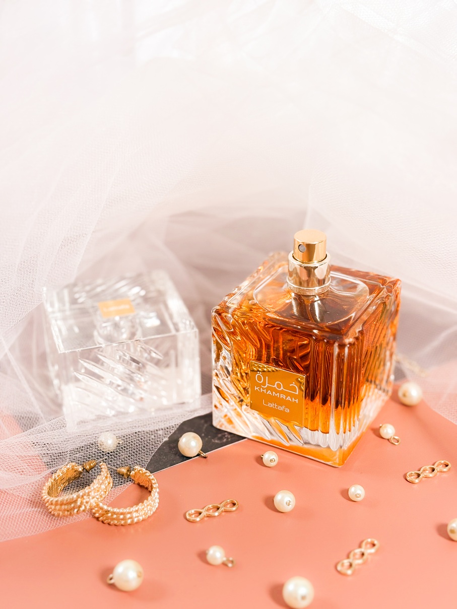 Le parfum Lattafa Khamrah ouvert, avec son bouchon posé à côté du flacon facetté ambré, sur un fond délicat avec voile en tulle, perles et bijoux dorés.