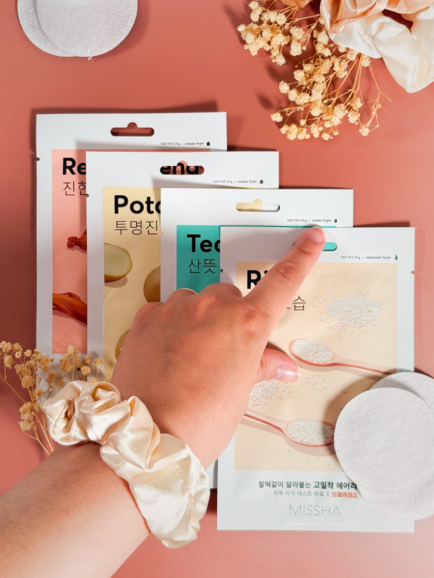 Main pointant les masques en tissu Missha Airy Fit Mask disposés sur un fond rose délicat avec cotons, fleurs séchées et chouchou satiné.