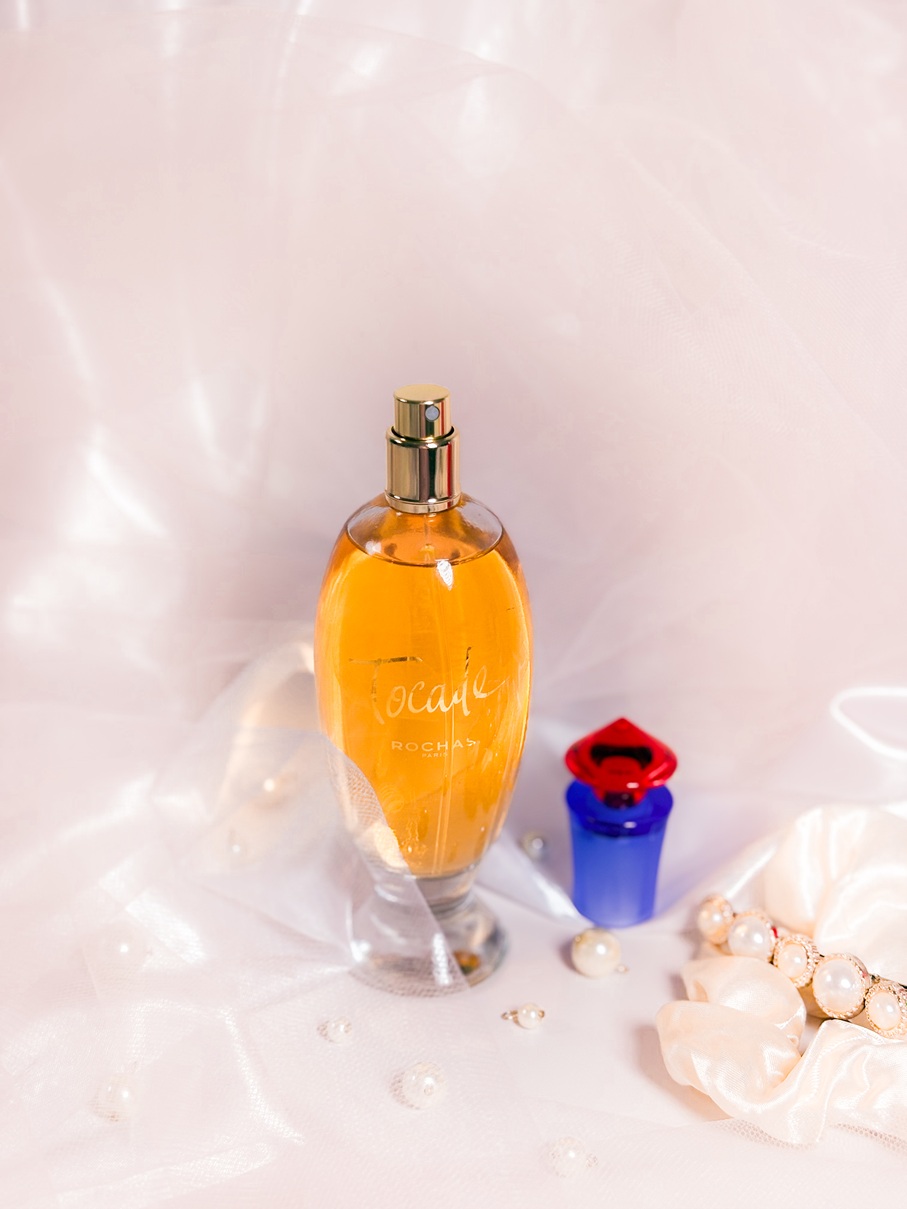 Le parfum Rochas Tocade ouvert, avec son bouchon bicolore posé à côté du flacon ambré, sur un fond délicat avec tissu satiné, perles, chouchou satiné et barrette ornée de perles.