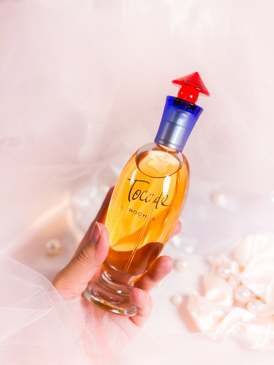 Main tenant le parfum Rochas Tocade dans son flacon ambré aux accents rouges et bleus, sur un fond délicat avec voile en tulle, perles, chouchou satiné et barrette ornée de perles.