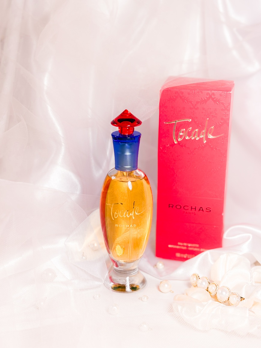 Le parfum Rochas Tocade et sa boîte rose posés sur un fond délicat avec voile en tulle, perles, chouchou satiné et barrette ornée de perles.