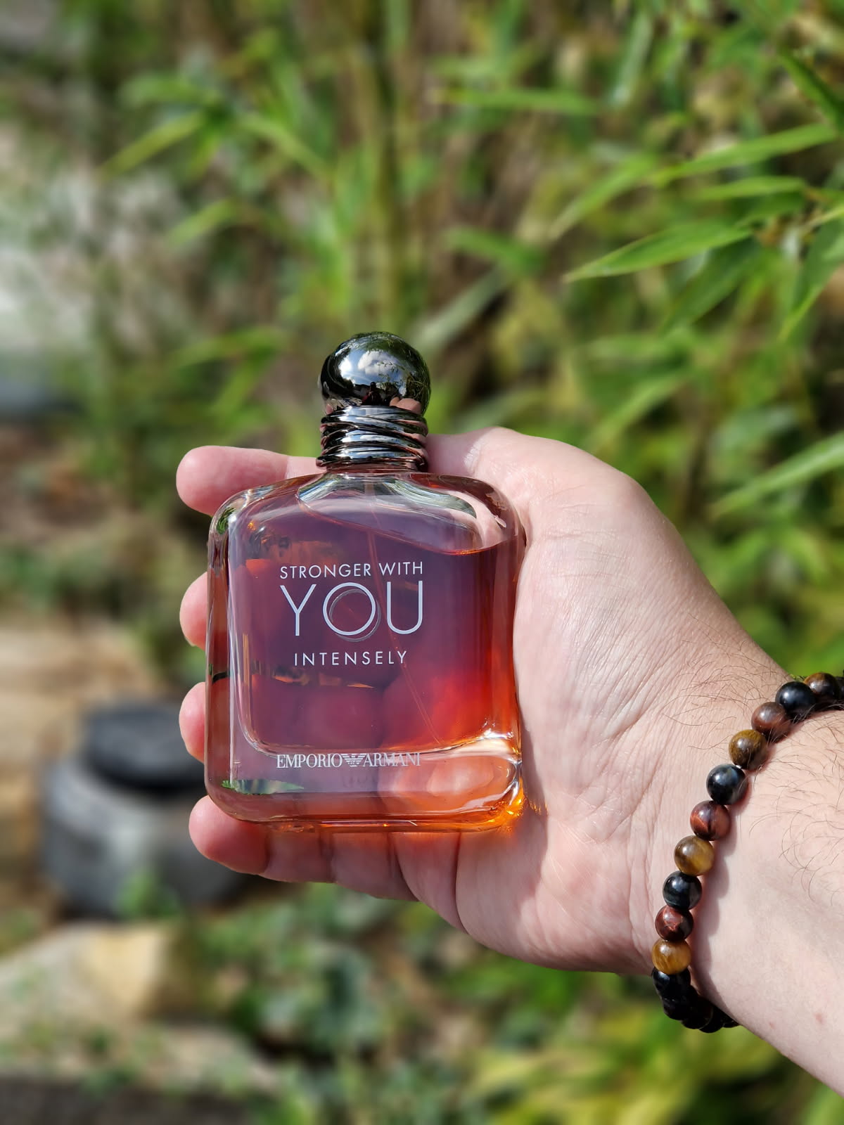 Main tenant le parfum Emporio Armani Stronger With You Intensely dans son flacon ambré, sur un fond naturel aux tons verts dans une mise en scène élégante et masculine.