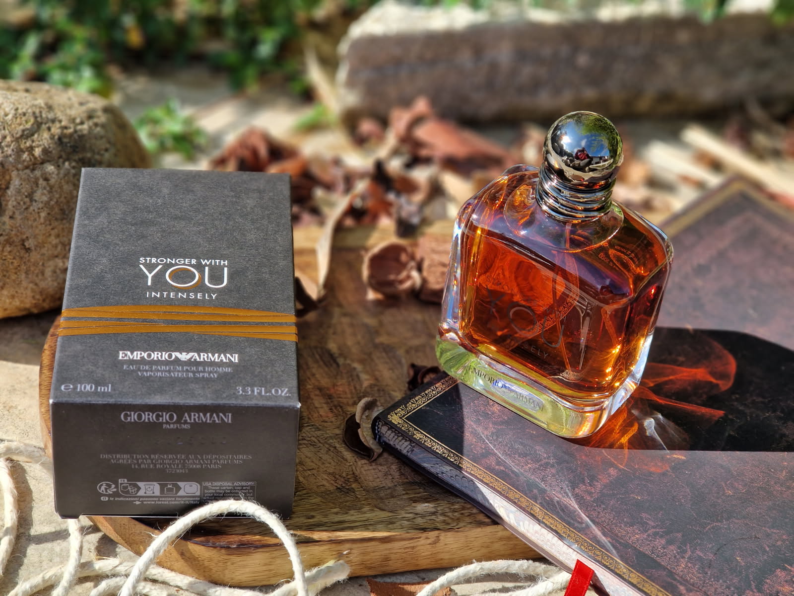 Le parfum Emporio Armani Stronger With You Intensely et sa boîte noire posés sur une planche en bois, dans une mise en scène chaleureuse avec carnet, corde, épices et décor naturel en arrière-plan.