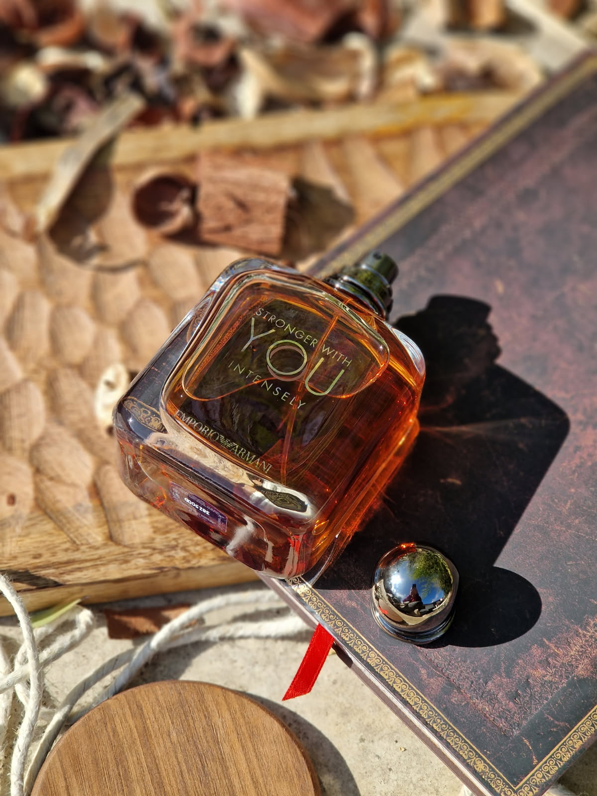 Le parfum Emporio Armani Stronger With You Intensely ouvert, avec son bouchon sphérique posé à côté du flacon ambré, sur une planche en bois avec carnet, corde et épices, dans une mise en scène chaleureuse aux accents masculins et raffinés.