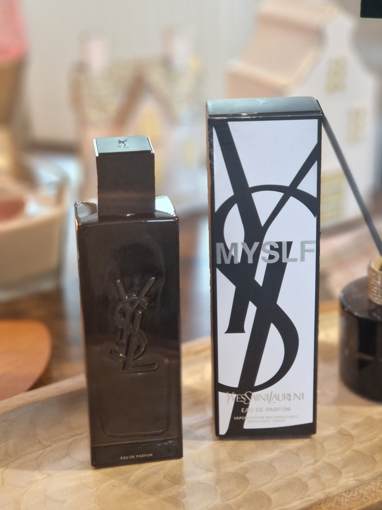 Le parfum Yves Saint Laurent MYSLF dans son flacon noir laqué, posé à côté de sa boîte blanche et noire sur une table, avec un diffuseur, des petites maisons lumineuses et une ambiance chaleureuse en arrière-plan.