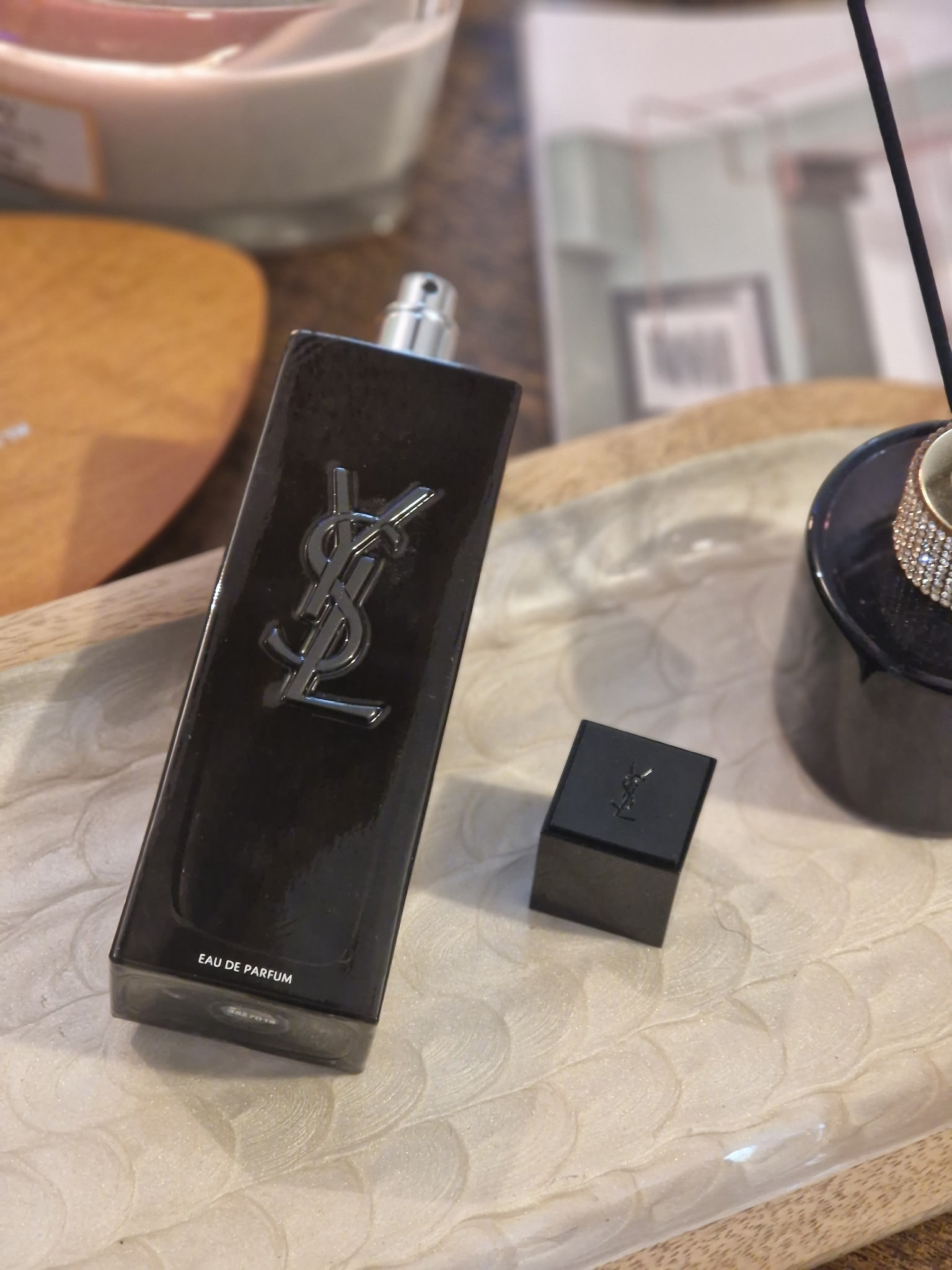 Le parfum Yves Saint Laurent MYSLF ouvert, avec son bouchon carré noir posé à côté du flacon noir laqué, sur un plateau aux côtés d’une bougie allumée, d’un diffuseur et d’un magazine, dans une ambiance chaleureuse et cocooning.