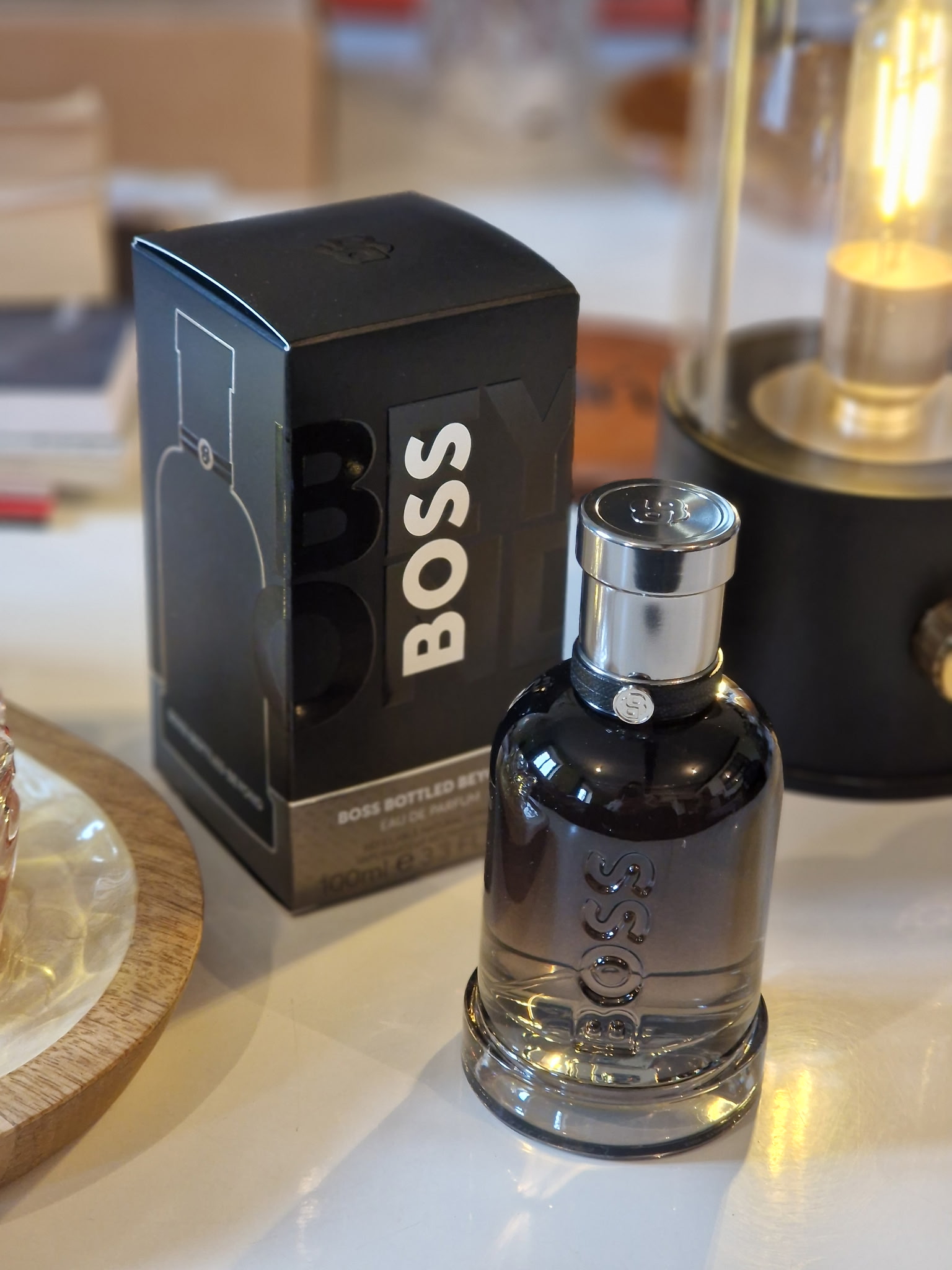 Le parfum Hugo Boss Boss Bottled Beyond dans son flacon noir fumé, posé à côté de sa boîte noire sur une table, avec une lampe décorative allumée en arrière-plan dans une ambiance élégante et chaleureuse.