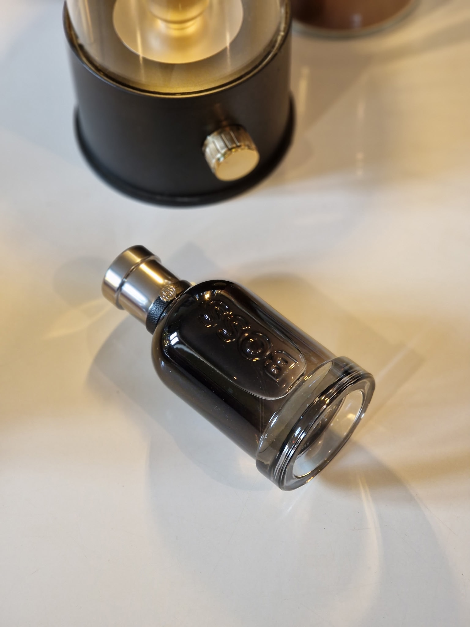 Le parfum Hugo Boss Boss Bottled Beyond posé seul, dans son flacon noir fumé couché sur une table, avec une lampe décorative allumée en arrière-plan, dans une ambiance élégante et chaleureuse.