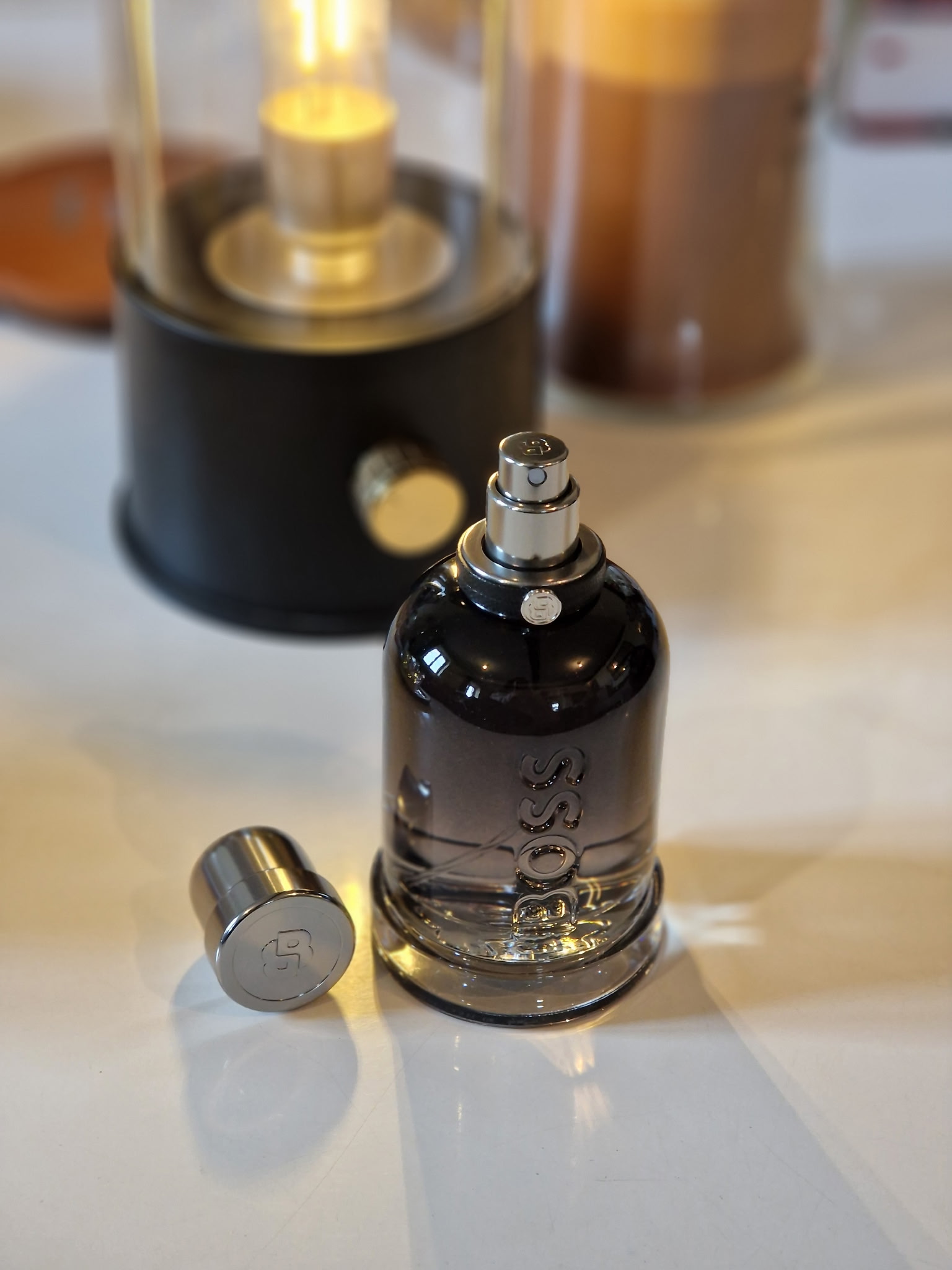 Le parfum Hugo Boss Boss Bottled Beyond ouvert, avec son bouchon argenté posé à côté du flacon noir fumé, sur une table avec une lampe décorative allumée en arrière-plan, dans une ambiance élégante et chaleureuse.