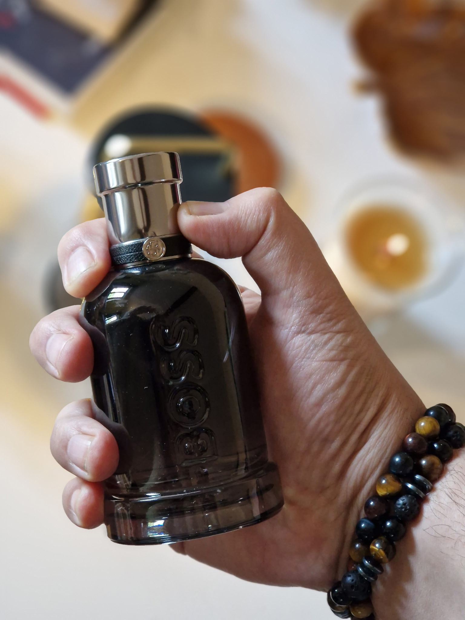 Main tenant le parfum Hugo Boss Boss Bottled Beyond dans son flacon noir fumé, sur un fond intérieur flou avec une bougie allumée et des éléments décoratifs, dans une ambiance élégante et chaleureuse.